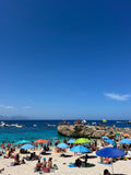 Beach scene with people, umbrellas, and boats under a clear blue sky.