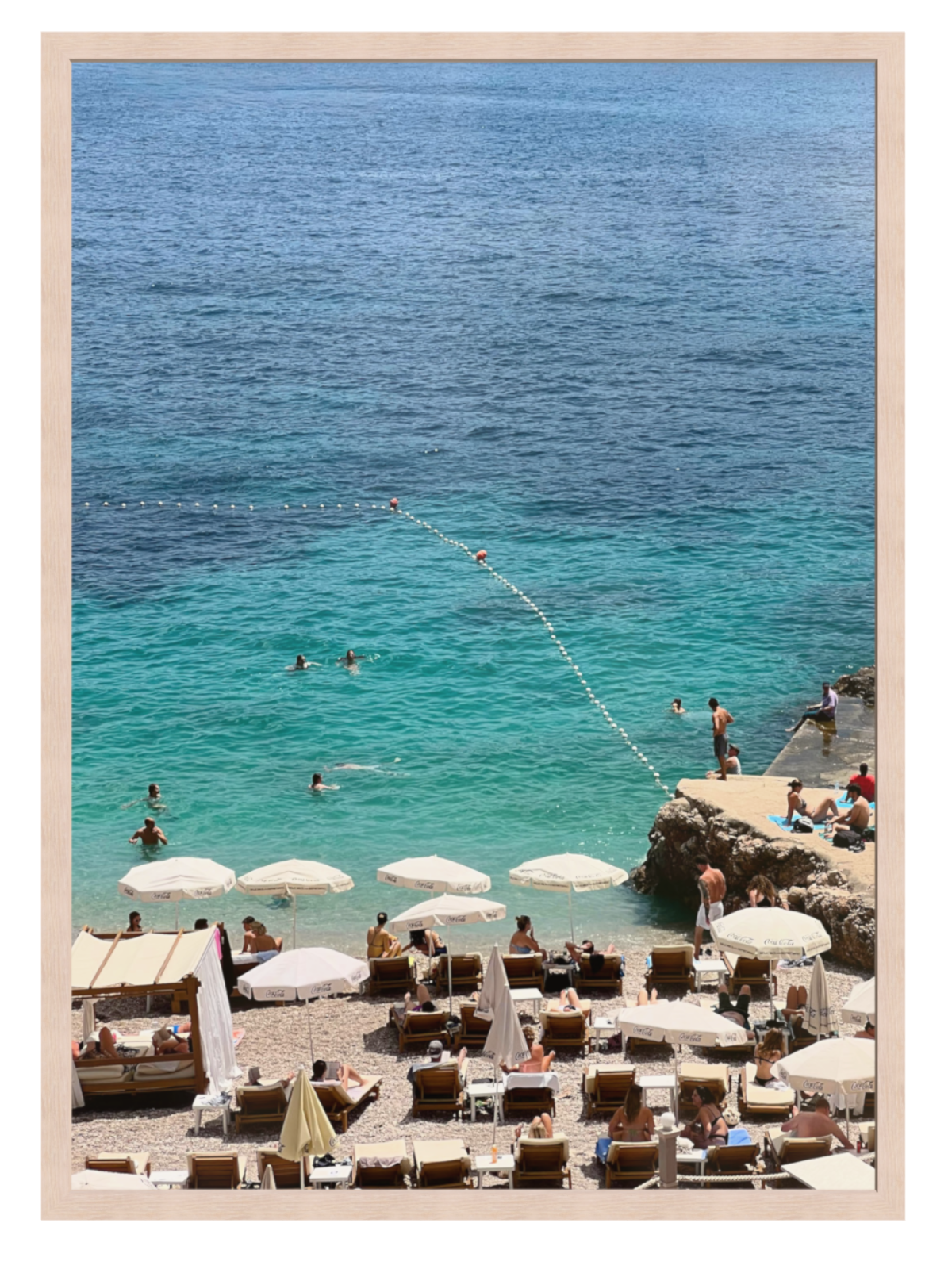 Beach scene with people, umbrellas, and lounge chairs near a body of water.Light oak frame