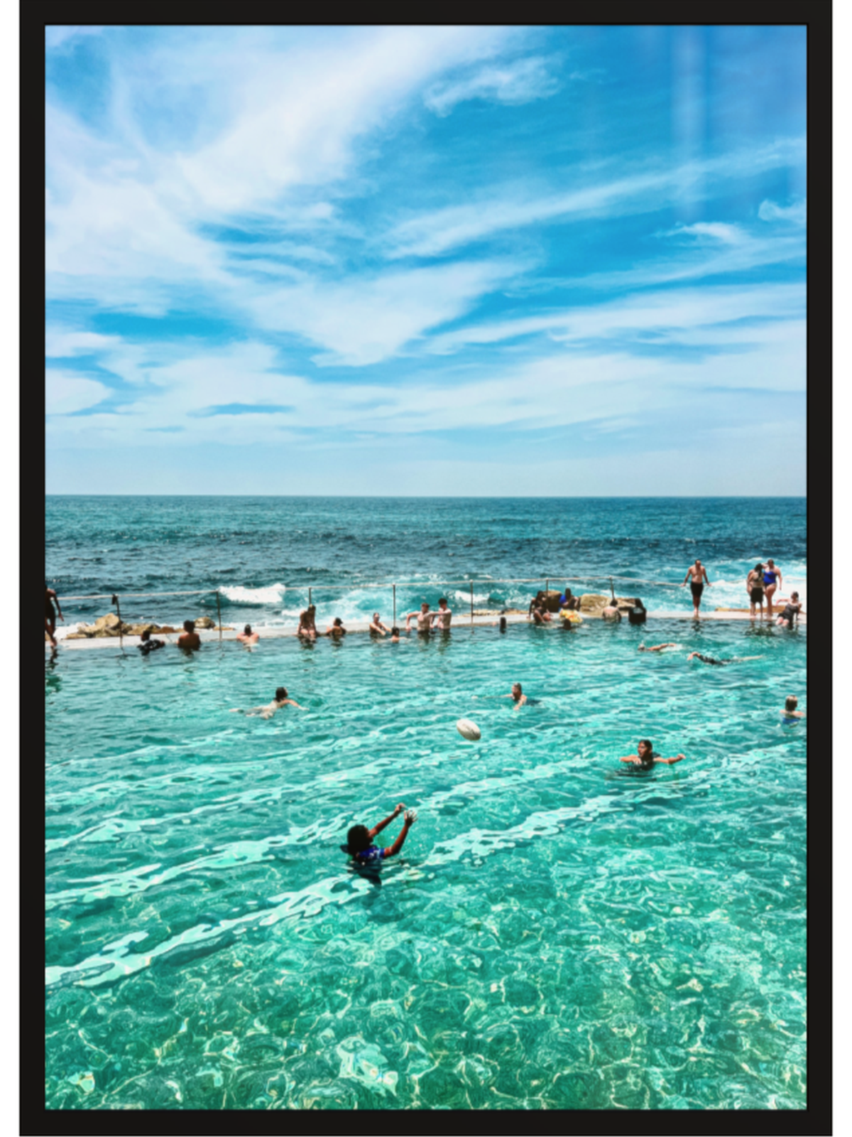 People swimming in a pool with ocean view under a blue sky.