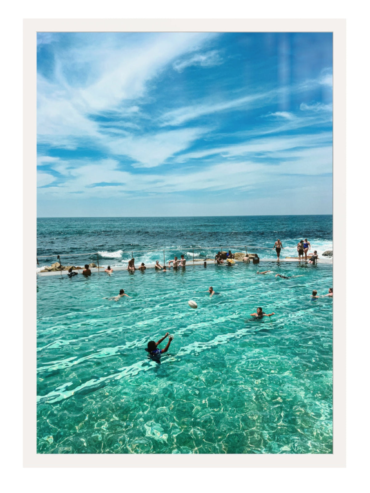 People swimming in a pool with ocean view under a blue sky.