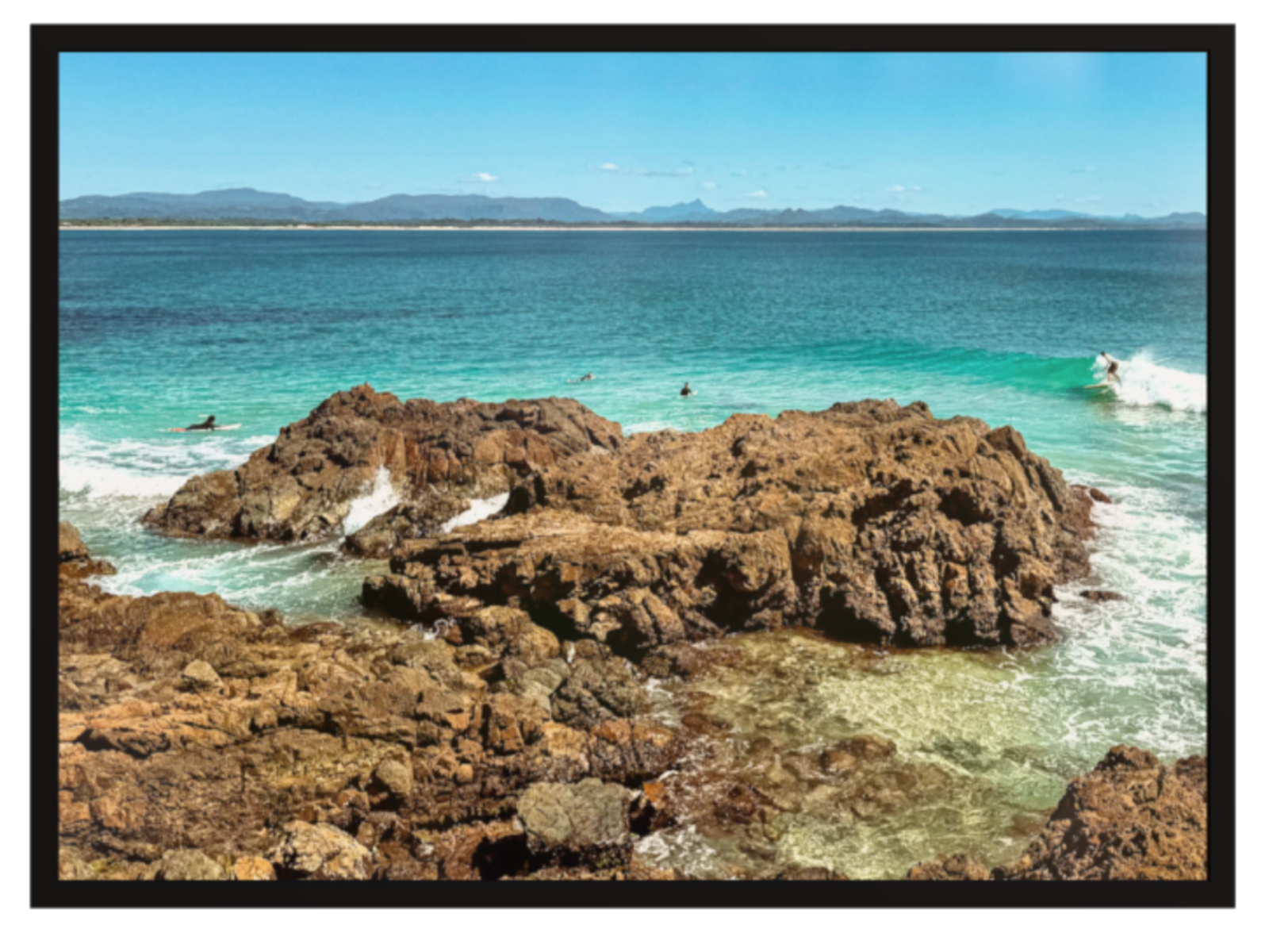Rocks by the ocean with clear blue water and distant mountains.