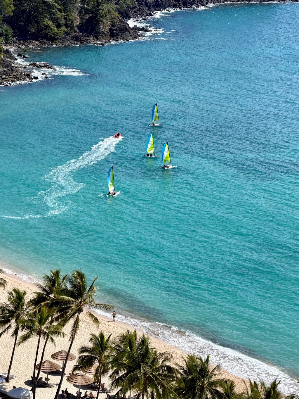 Aerial view of a tropical beach with turquoise water, several windsurfers on the sea, rocky shoreline, and palm trees along the sandy shore.