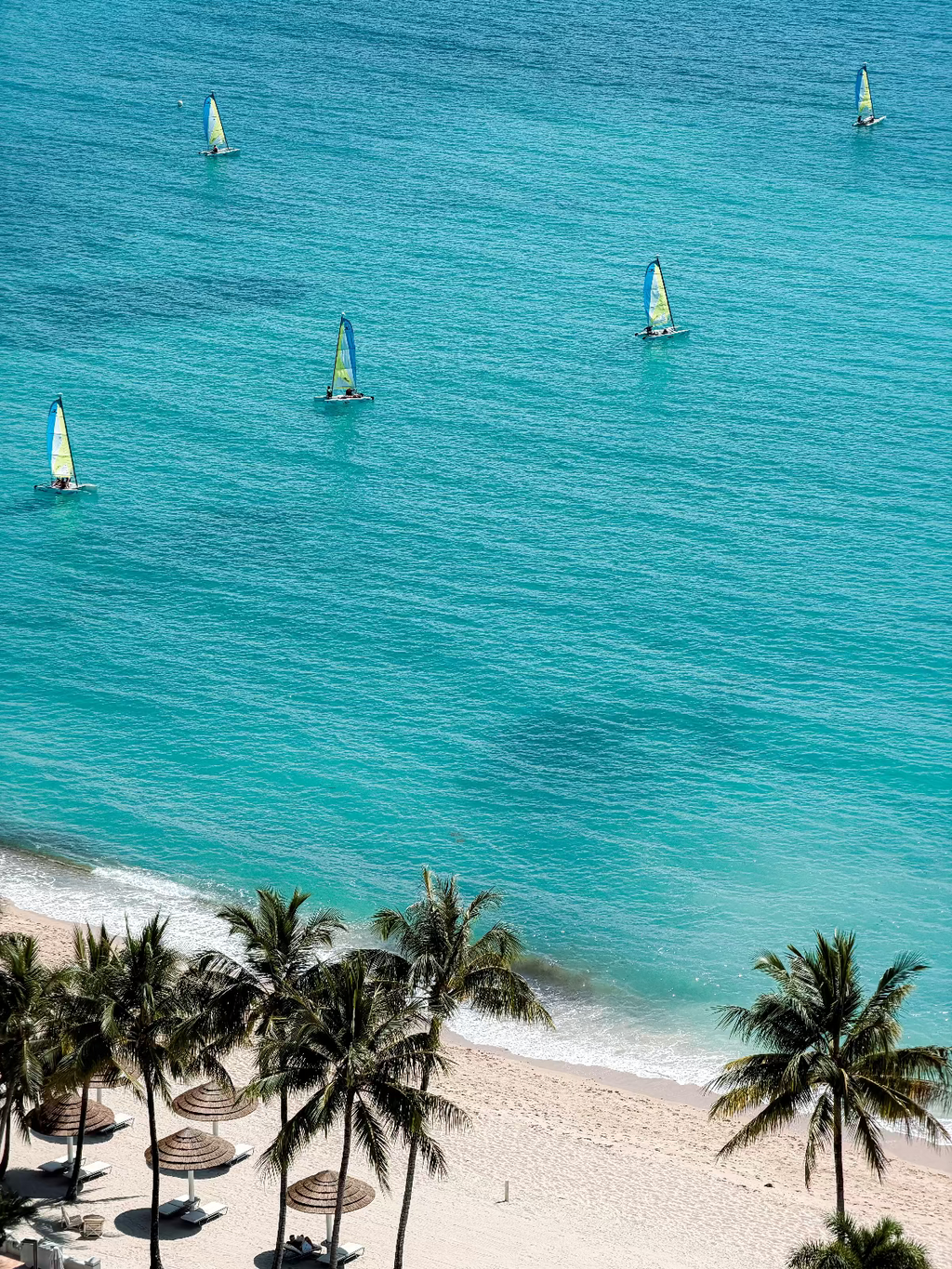 Aerial view of a tropical beach with turquoise ocean water, several sailboats on the sea, white sand, and palm trees along the shoreline.