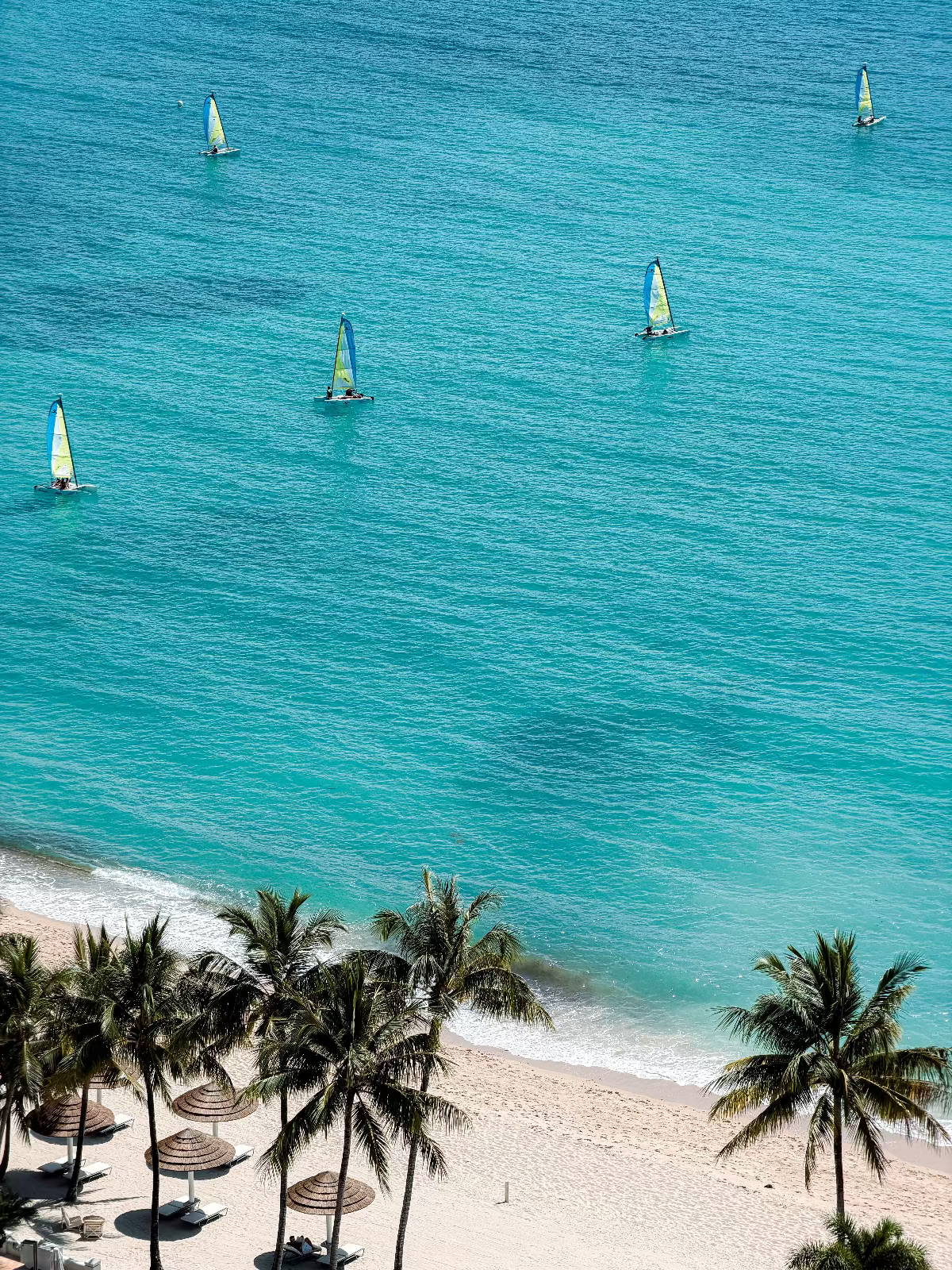 Aerial view of a tropical beach with turquoise ocean water, several sailboats on the sea, white sand, and palm trees along the shoreline.