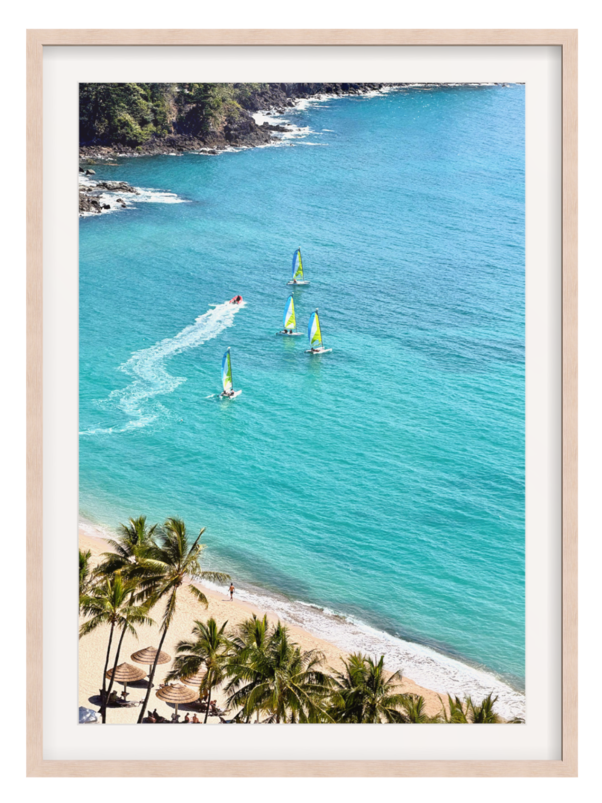 A framed art print featuring a scenic aerial photograph of a tropical beach with turquoise ocean water, palm trees, and windsurfers, set in a light wood frame with a white mat.
