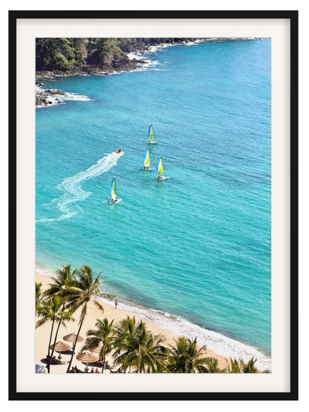 Framed wall art print showing a coastal beach scene with turquoise water, sailboats, a speedboat leaving a white wake, sandy shore, and palm trees, presented in a black frame with white matting.