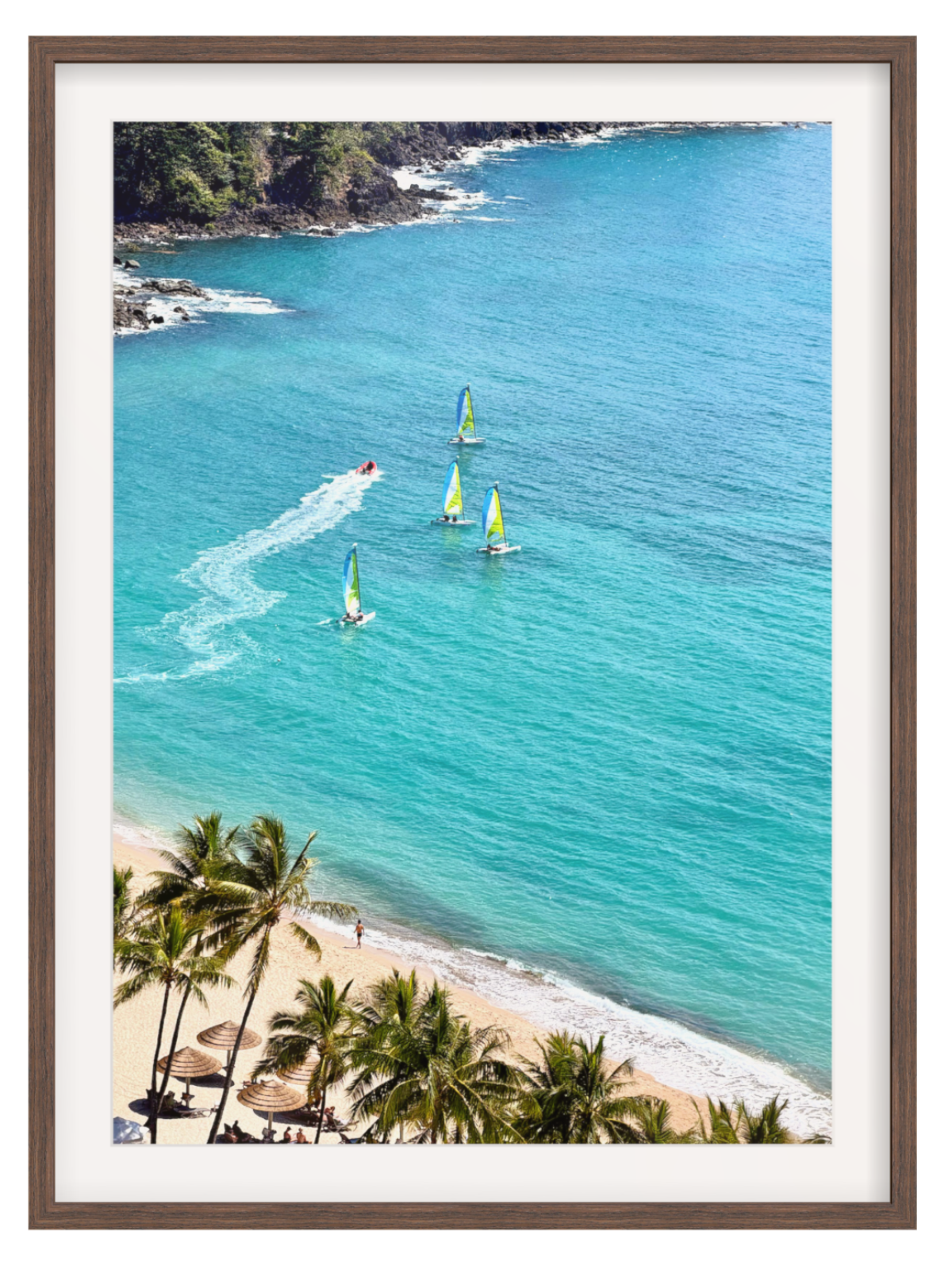 Framed wall art print showing a tropical coastal scene with turquoise ocean water, sailboats, a small motorboat leaving a white wake, sandy beach, palm trees, and rocky shoreline. The print is presented with a white mat and brown frame against a light gray background.