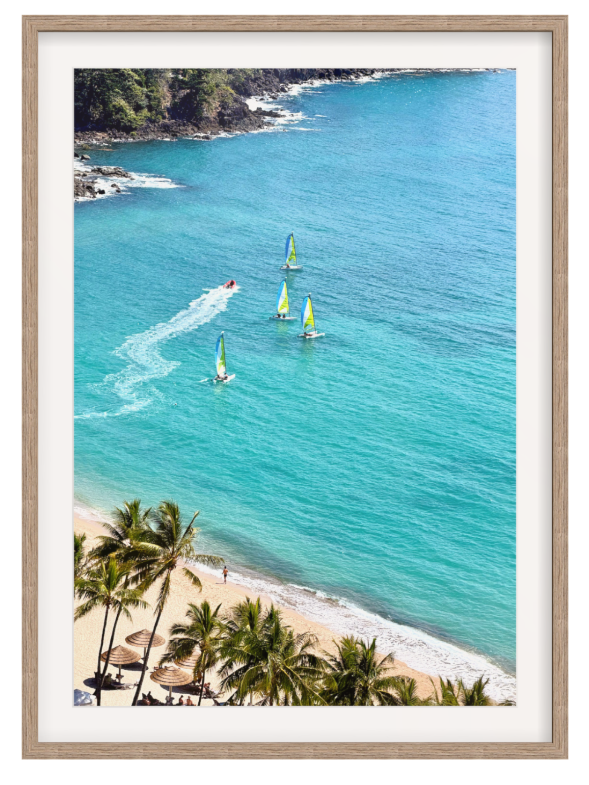 A framed coastal art print showing a tropical beach with palm trees, turquoise water, and several sailboats, presented in a light wood frame with a white mat.