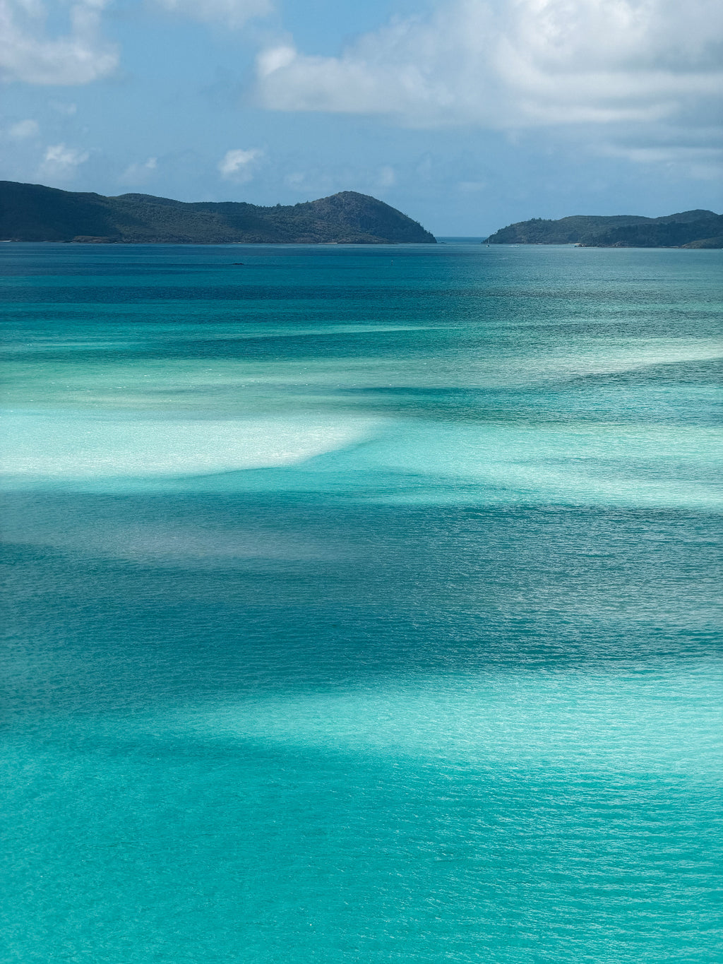 Scenic view of turquoise ocean water with shallow clear pools, distant dark green hills, and a blue sky with white clouds.