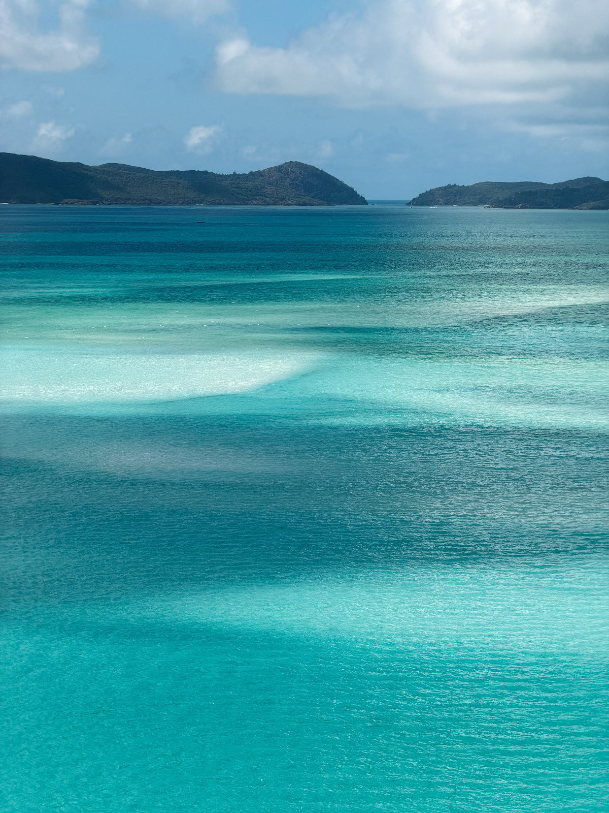 Scenic view of turquoise ocean water with shallow clear pools, distant dark green hills, and a blue sky with white clouds.
