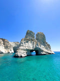 Rock formation with a natural arch in clear blue water under a bright blue sky.