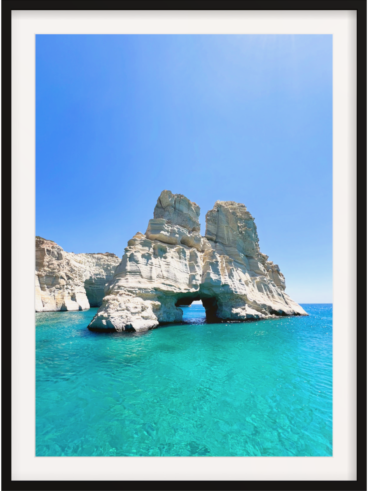 Rock formation with a natural arch in clear blue water under a bright blue sky.
