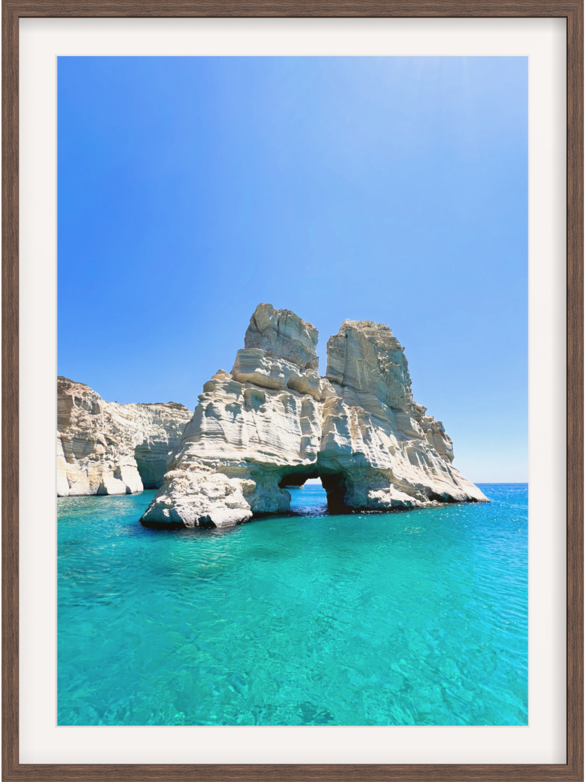 Rock formation with a natural arch in clear blue water under a bright blue sky.