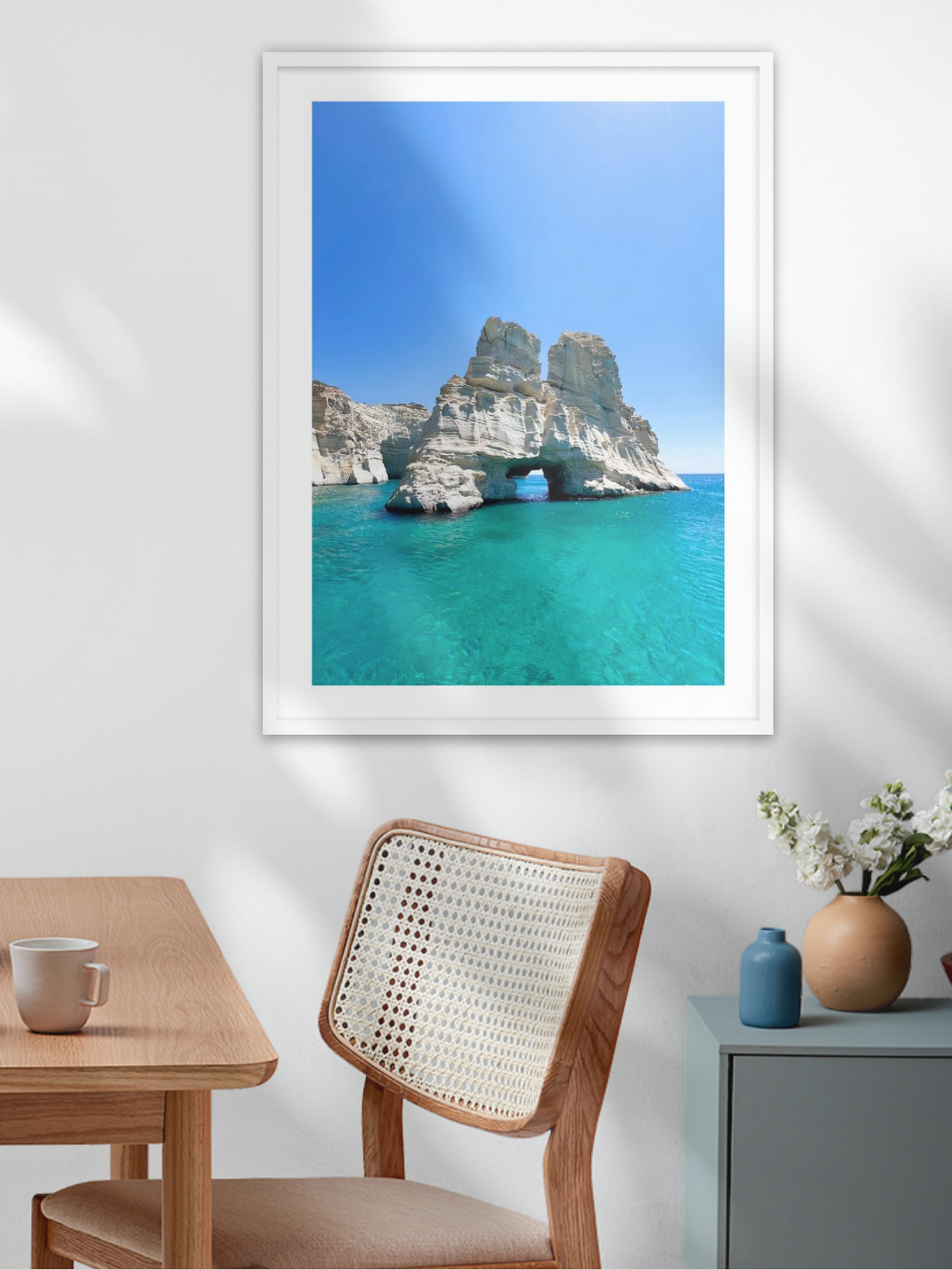 Framed artwork of a coastal scene with cliffs and turquoise water in a room with a table and chair.