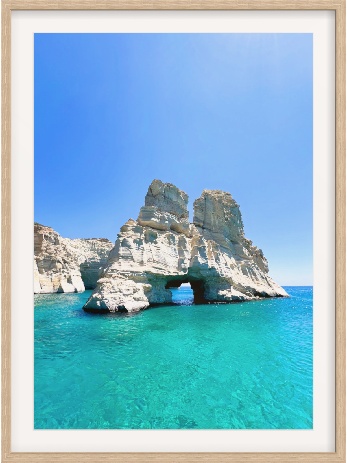 Rock formation with a natural arch in clear blue water under a bright blue sky.
