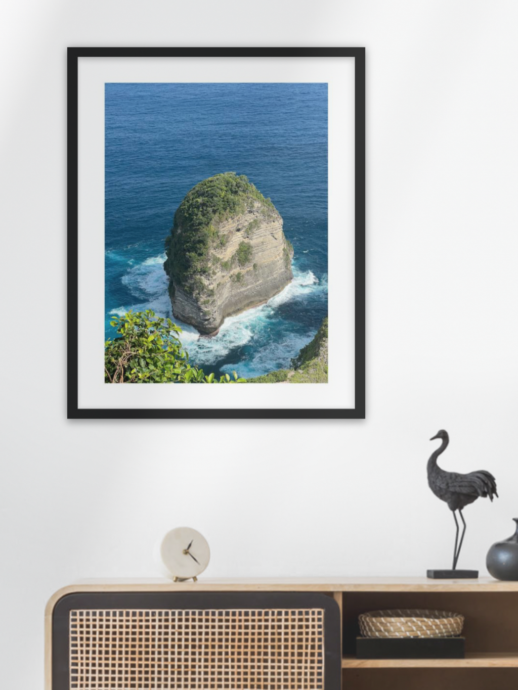 Framed photograph of a rocky island with ocean waves on a white wall above a wooden cabinet.