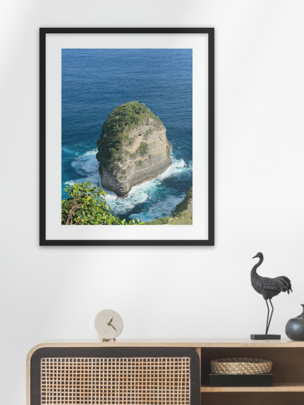 Framed photograph of a rocky island with ocean waves on a white wall above a wooden cabinet.