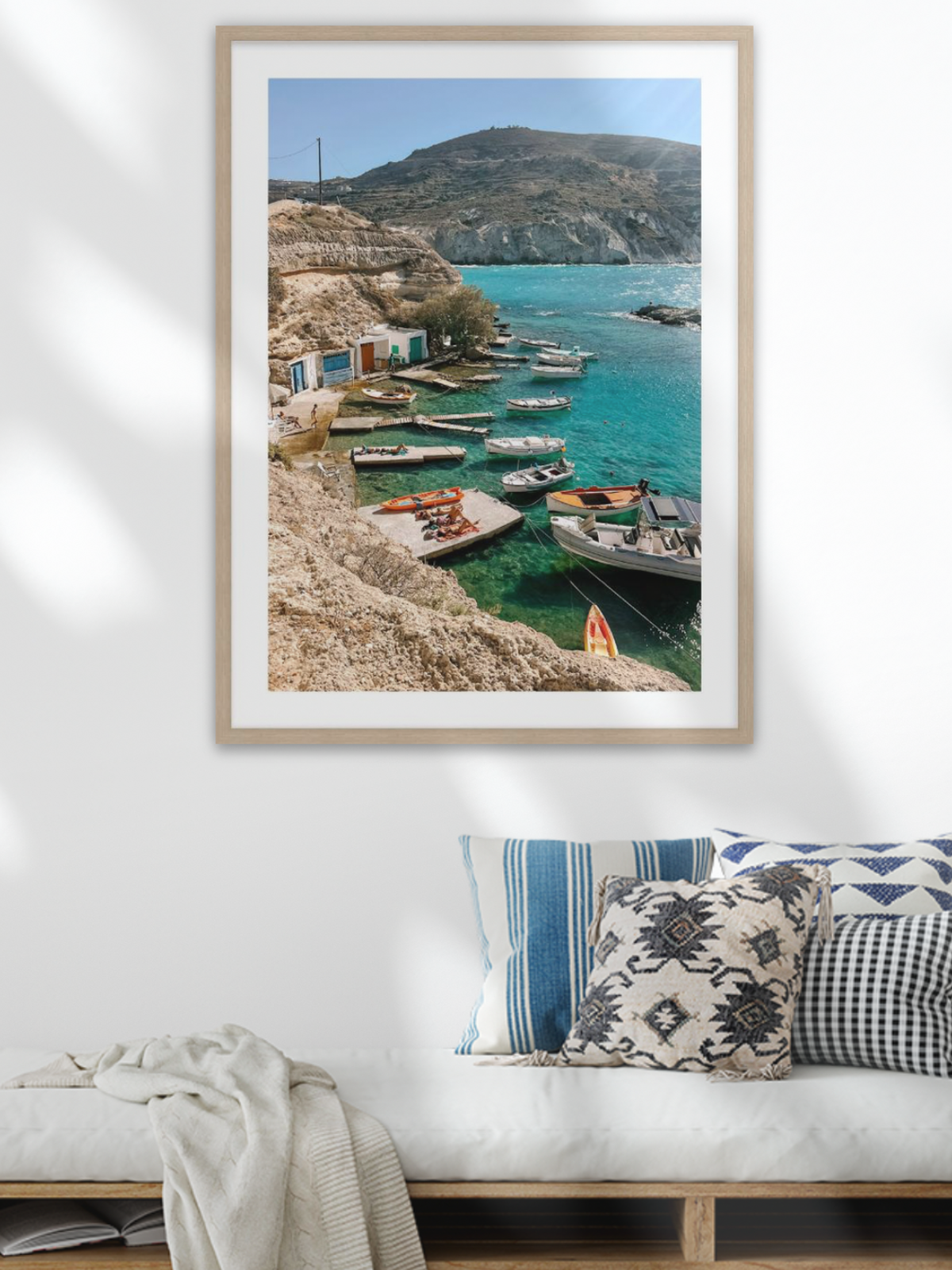 Framed artwork of a coastal scene with boats and mountains, displayed on a wall above a bench.