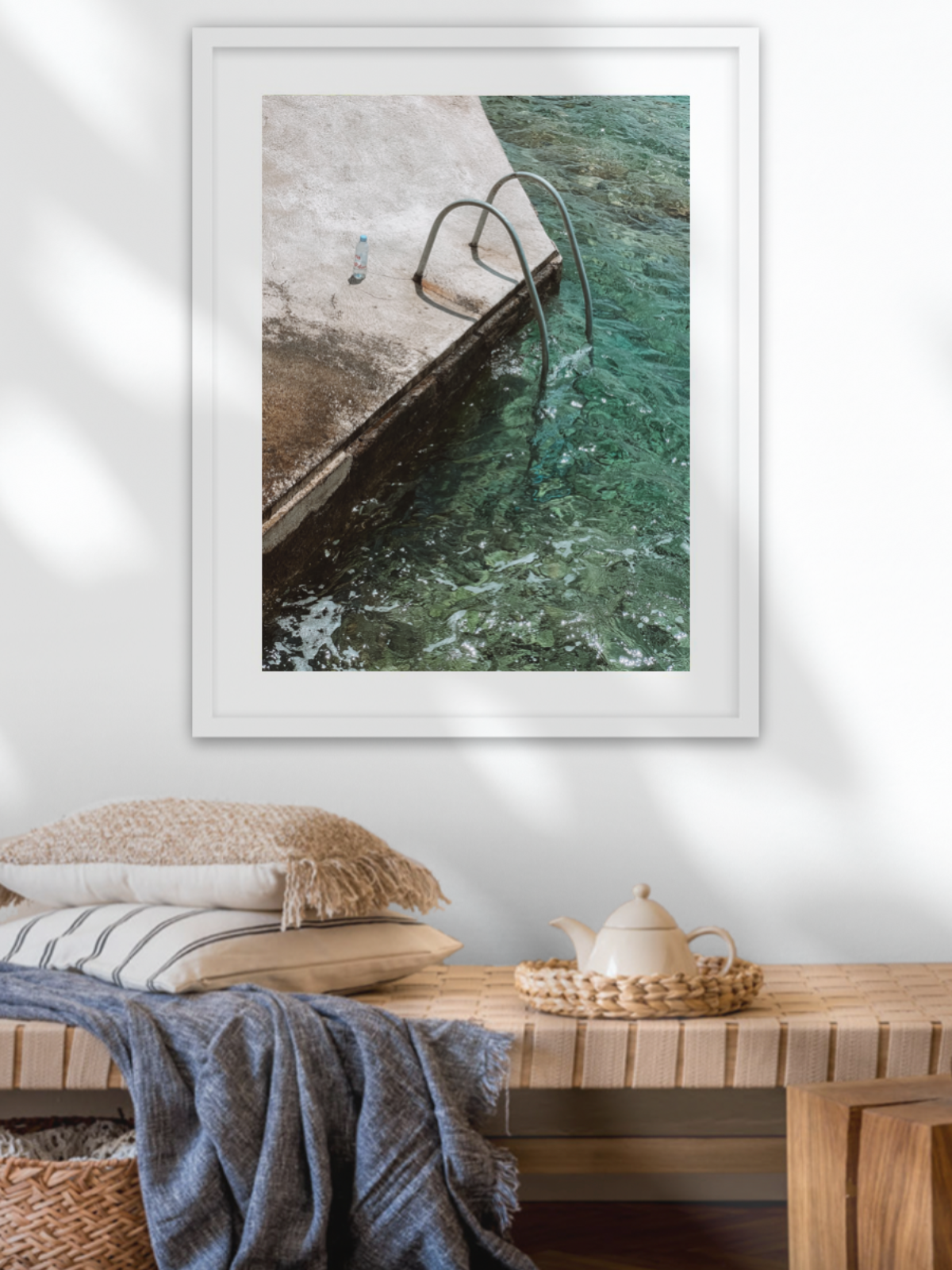 Framed artwork of a poolside scene on a wall above a wooden bench with pillows and decor.