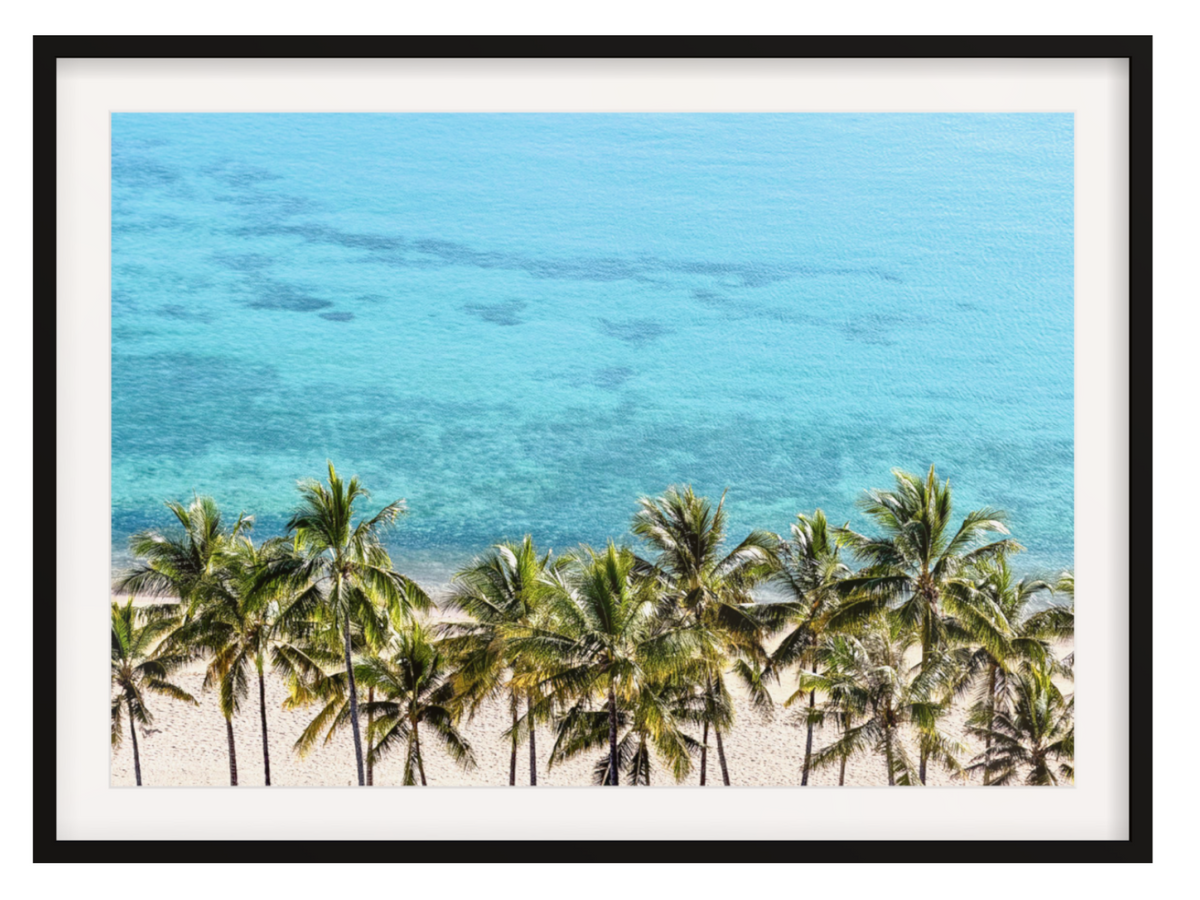 Framed wall art featuring a tropical beach scene with turquoise ocean water, a sandy shore, and a row of green palm trees, shown with a white mat and black frame.