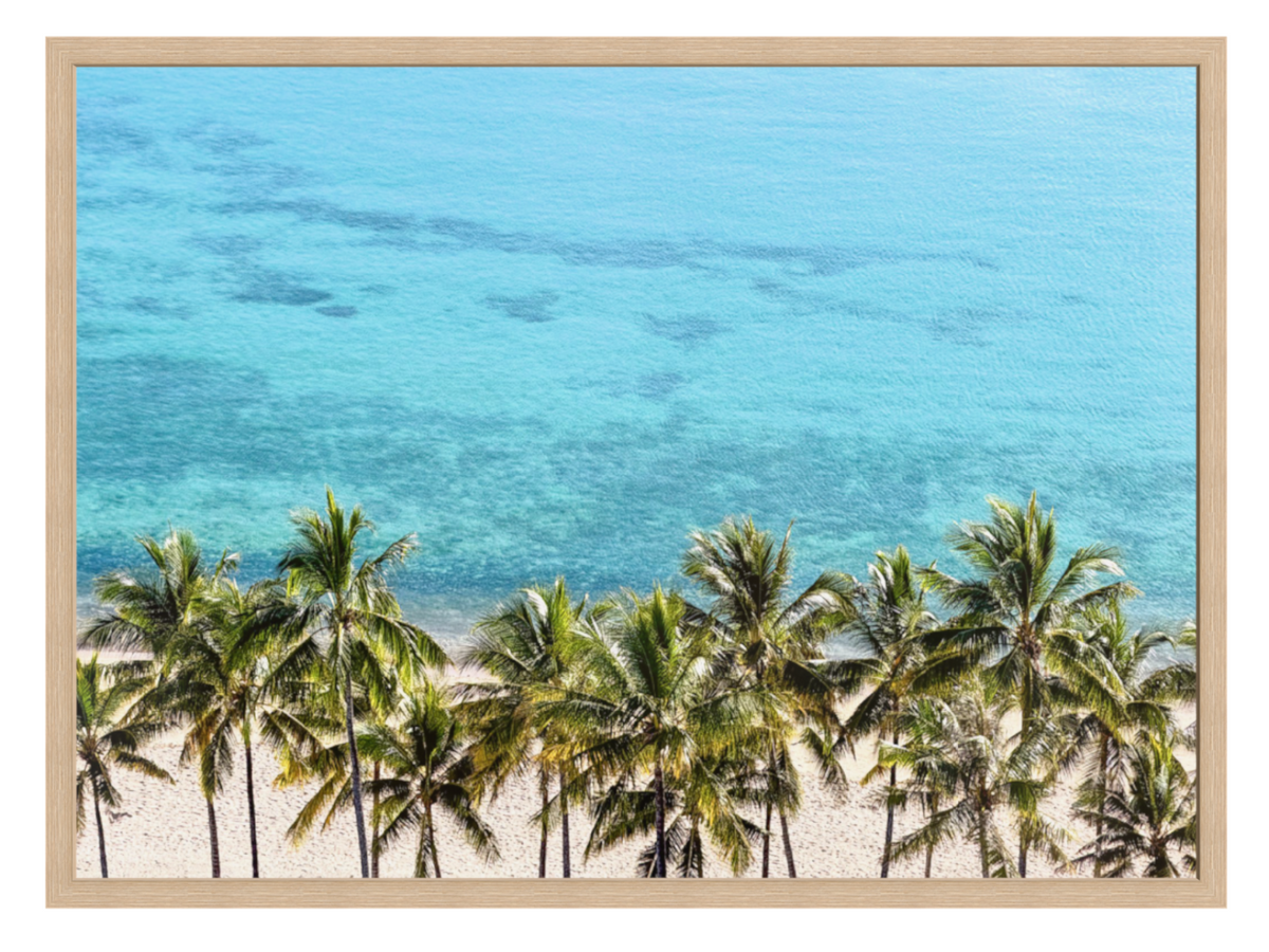 A framed photographic print of a tropical beach scene, featuring green palm trees on a sandy shore with clear turquoise ocean water under a light beige wood frame.