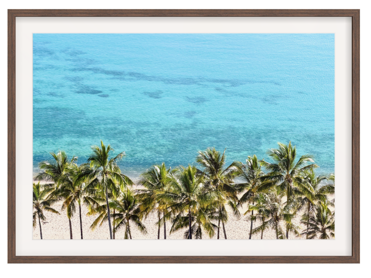 Framed wall art featuring a tropical beach scene with palm trees along a sandy shore and bright turquoise ocean water, shown with a white mat and brown frame on a white background.