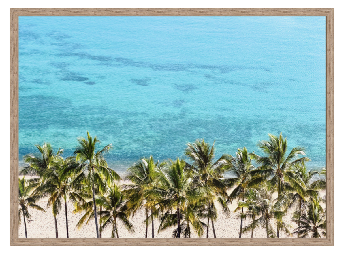 Framed wall art featuring a tropical beach scene with turquoise ocean water, white sand, and a row of green palm trees, shown in a light wood frame against a white background.