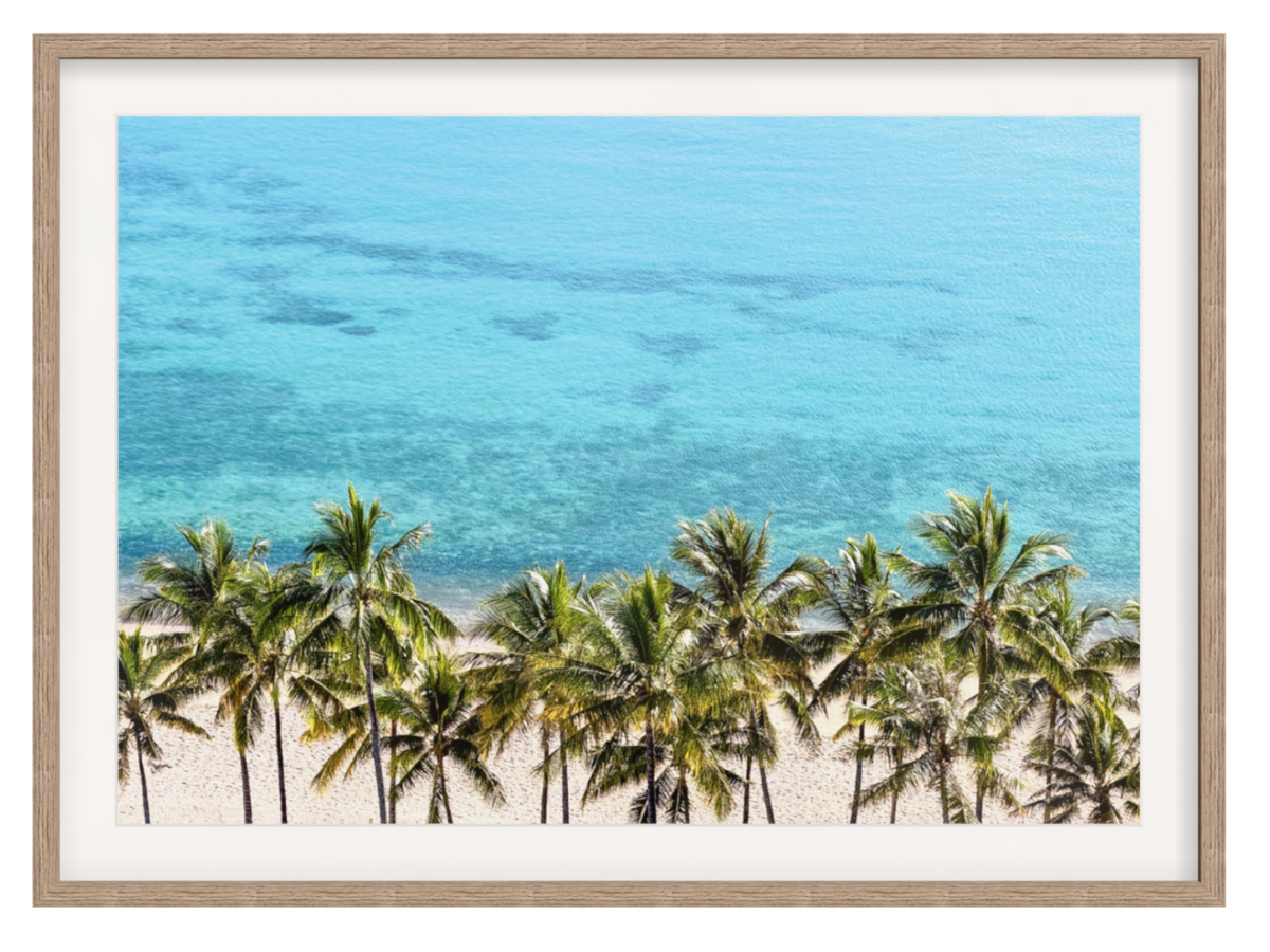 Framed wall art featuring a tropical beach scene with palm trees, white sand, and bright turquoise ocean water, shown in a light wood frame with a white mat on a white background.