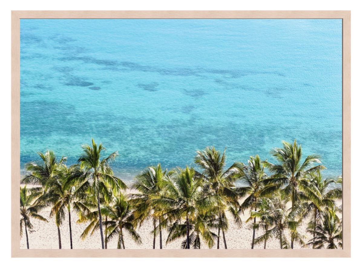 A framed tropical beach print featuring a row of green palm trees on a sandy shore overlooking clear turquoise ocean water, presented with a light wood frame.
