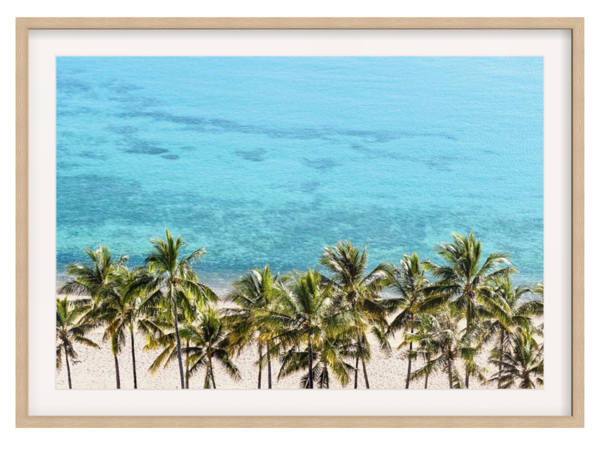 Framed wall art featuring a tropical beach scene with palm trees, white sand, and bright turquoise ocean water, shown in a light wood frame with a white mat on a white background.