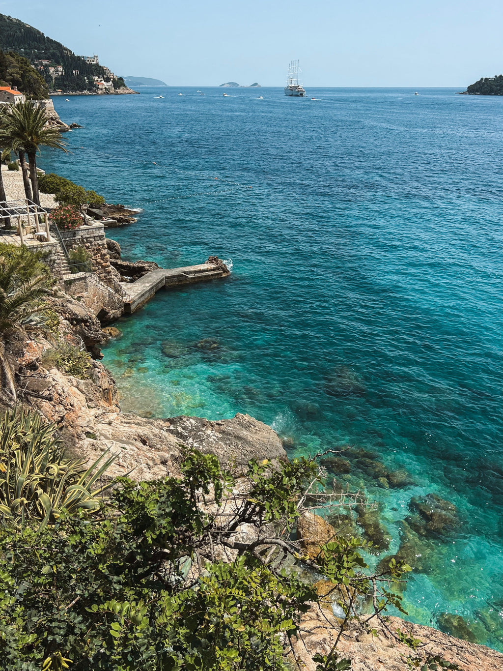 Coastal scene with clear blue water, rocky shore, and a sailboat in the distance.