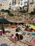 People sunbathing on a rocky beach with buildings and cliffs in the background