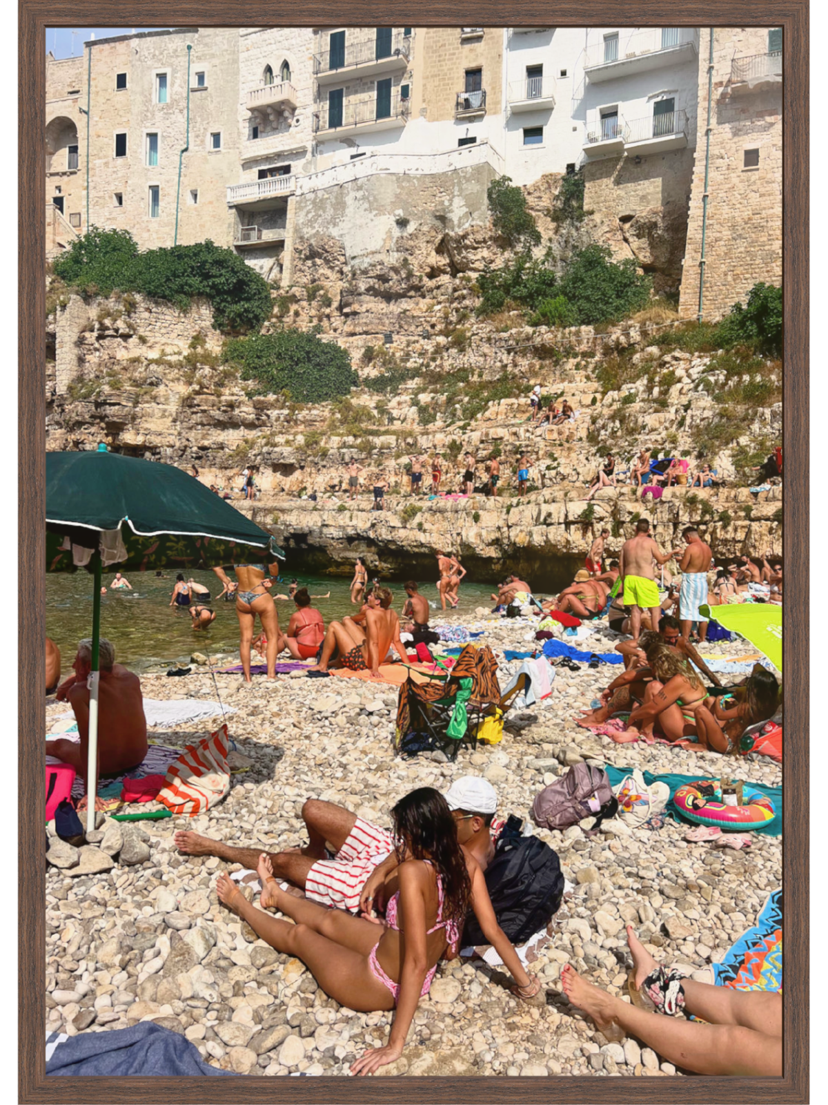 Beach scene with people sunbathing on a pebbly shore, buildings on a cliff in the background.