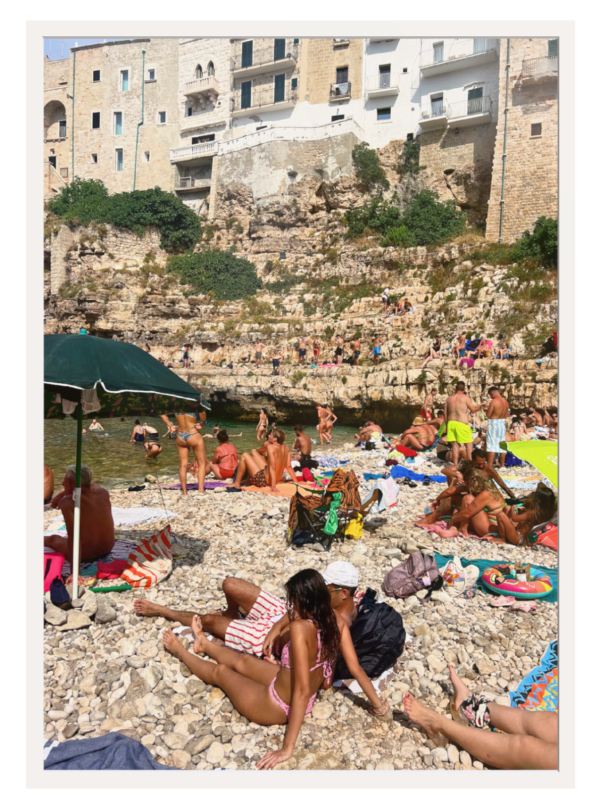 Beach scene with people sunbathing on a rocky shore with buildings in the background.