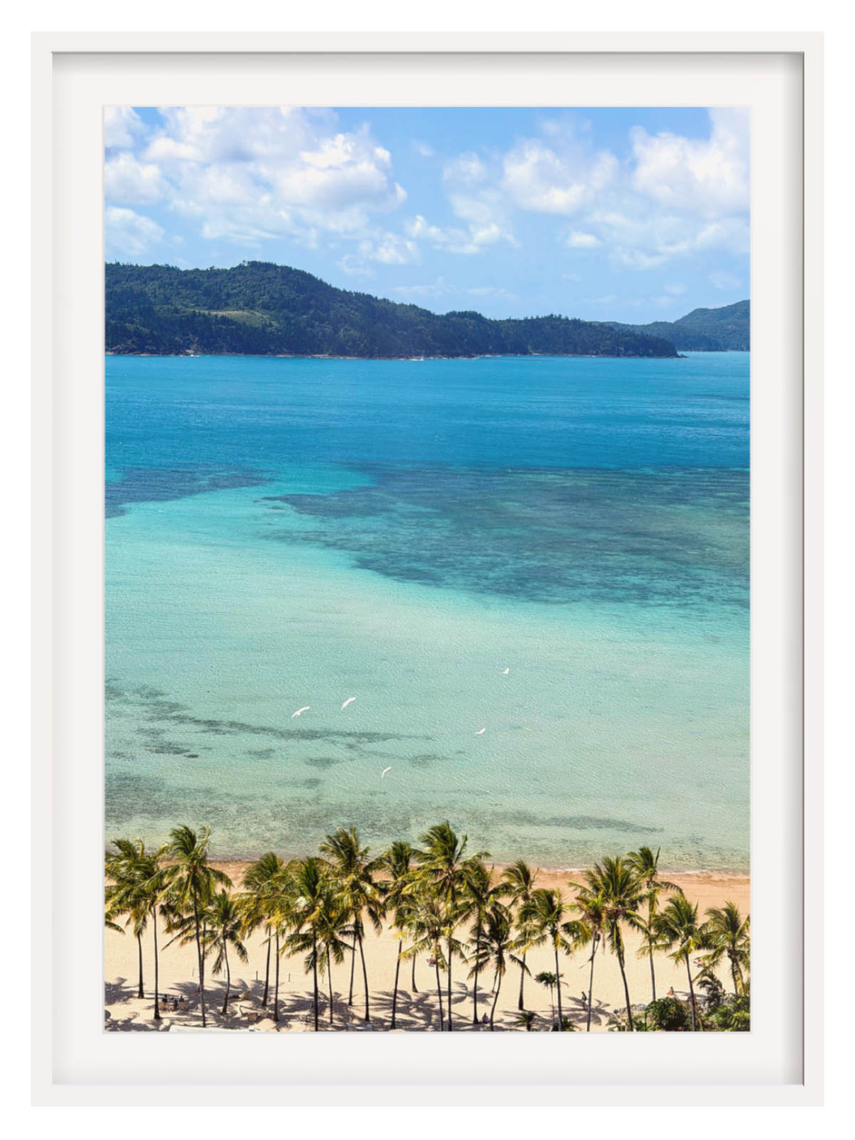 A framed art print showcasing a tropical beach scene with turquoise ocean water, a sandy shore lined with green palm trees, and distant green hills under a blue sky with white clouds.
