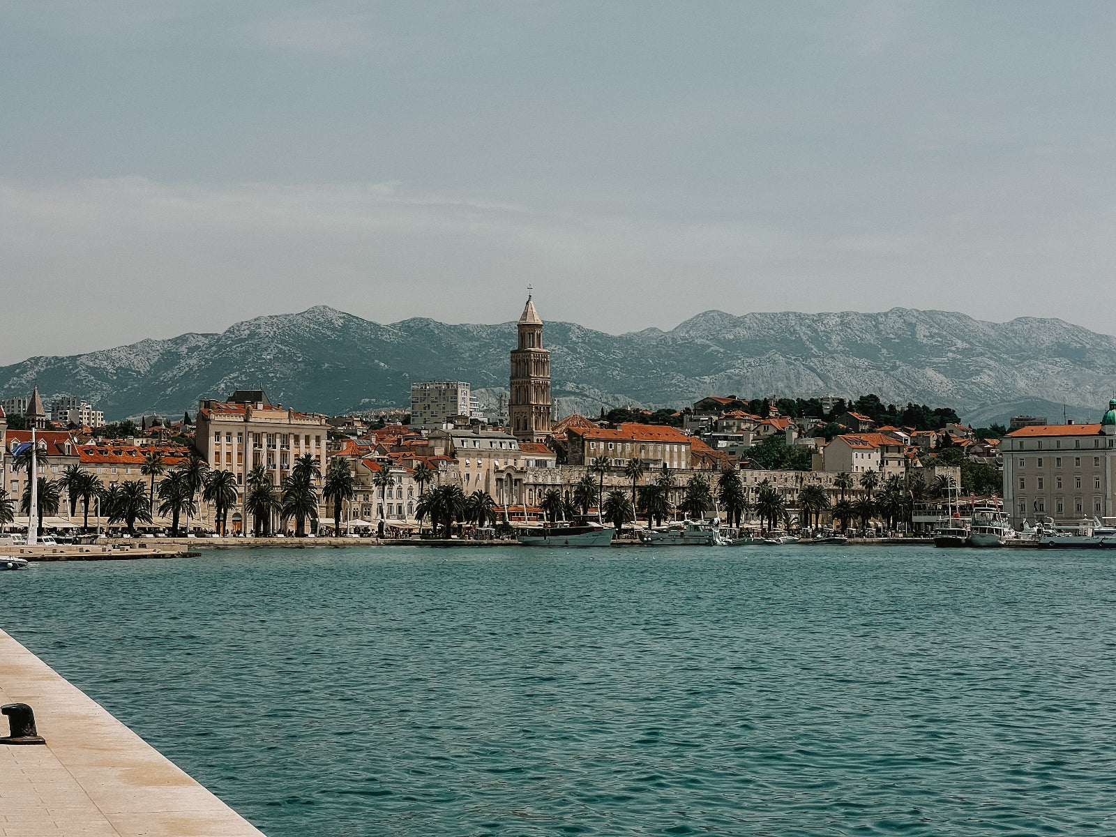 Cityscape with a prominent clock tower by a body of water, mountains in the background
