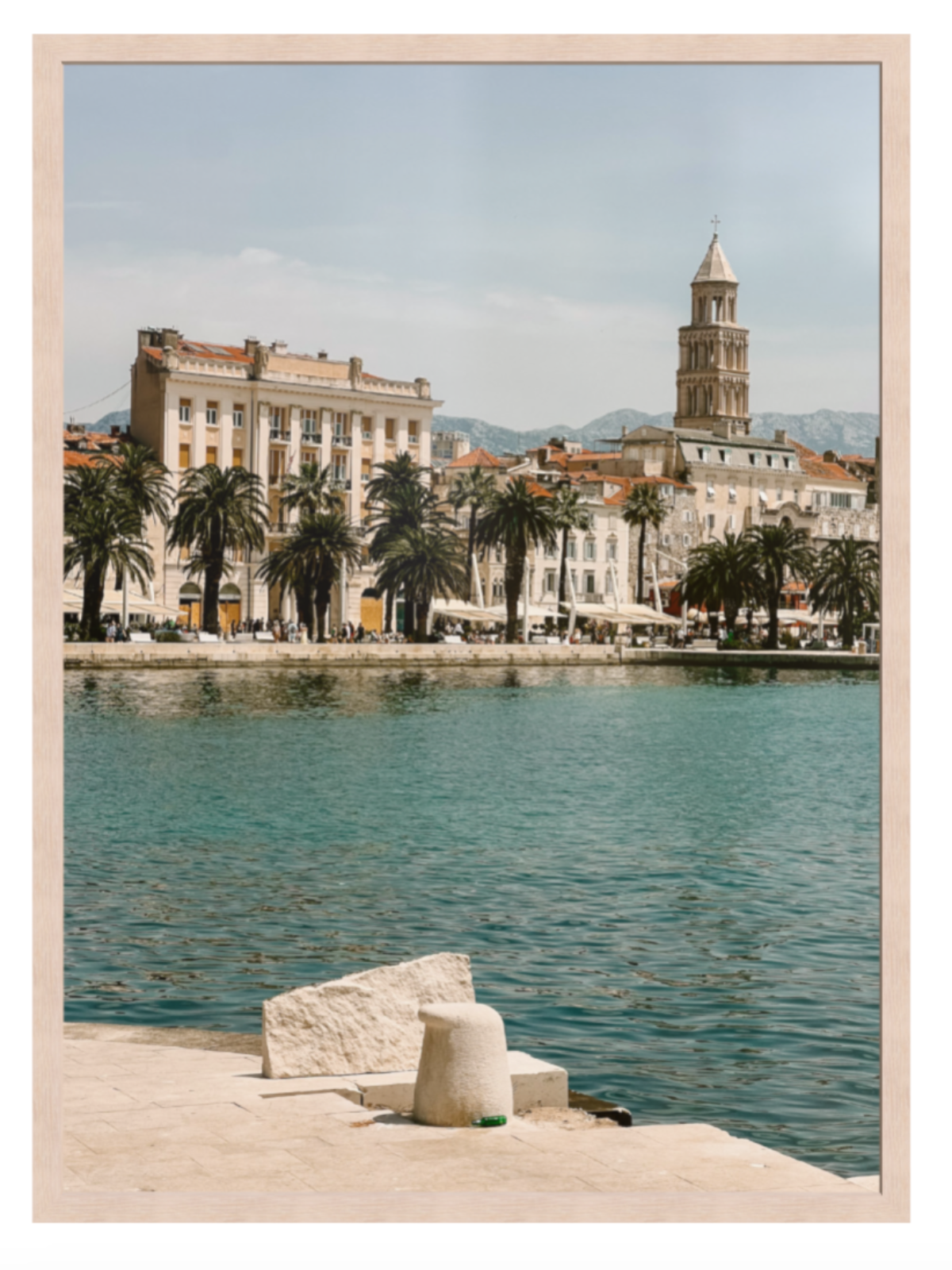 Cityscape with buildings and a clock tower by a waterfront, framed picture. Split light oak frame 