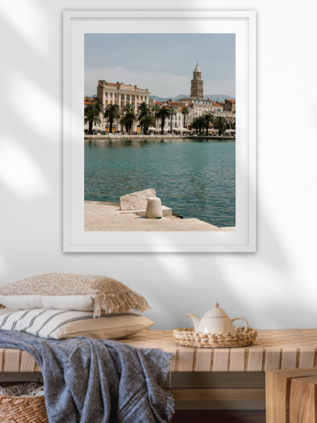 Framed artwork of a coastal scene with a cityscape on a wall above a wooden bench.
