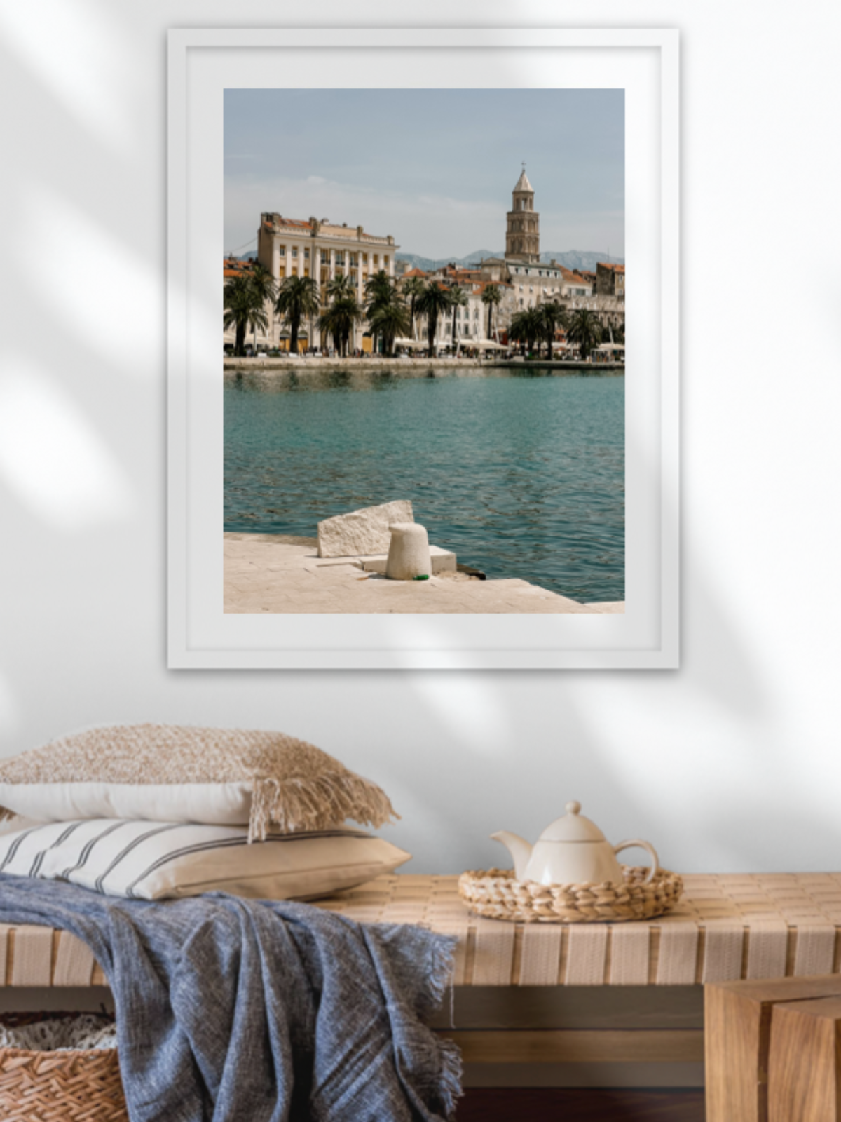 Framed artwork of a coastal scene with a cityscape on a wall above a wooden bench.