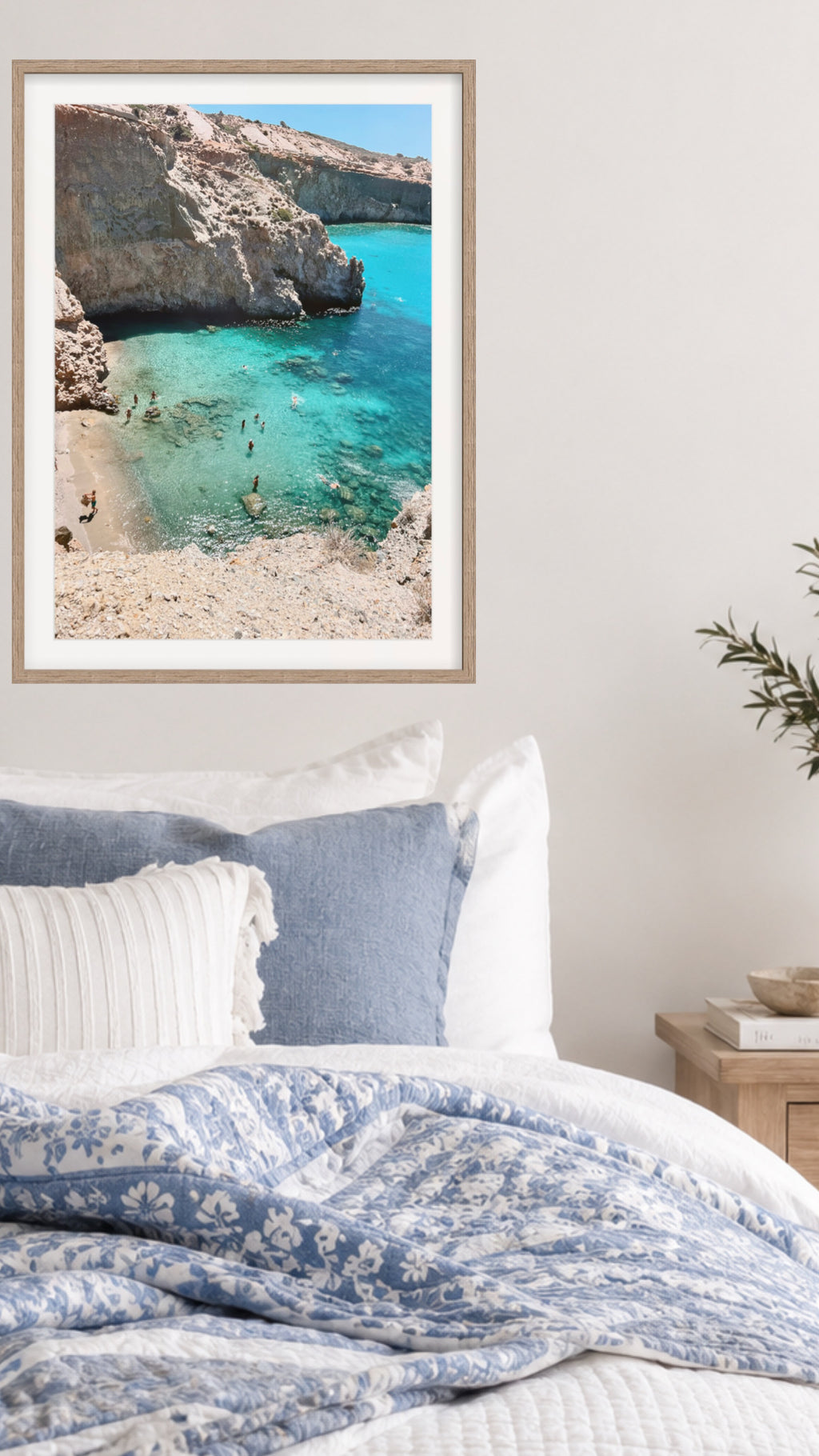 A framed art print of a scenic cove with turquoise water and rocky cliffs, styled on a light beige wall above a bed with blue and white bedding.