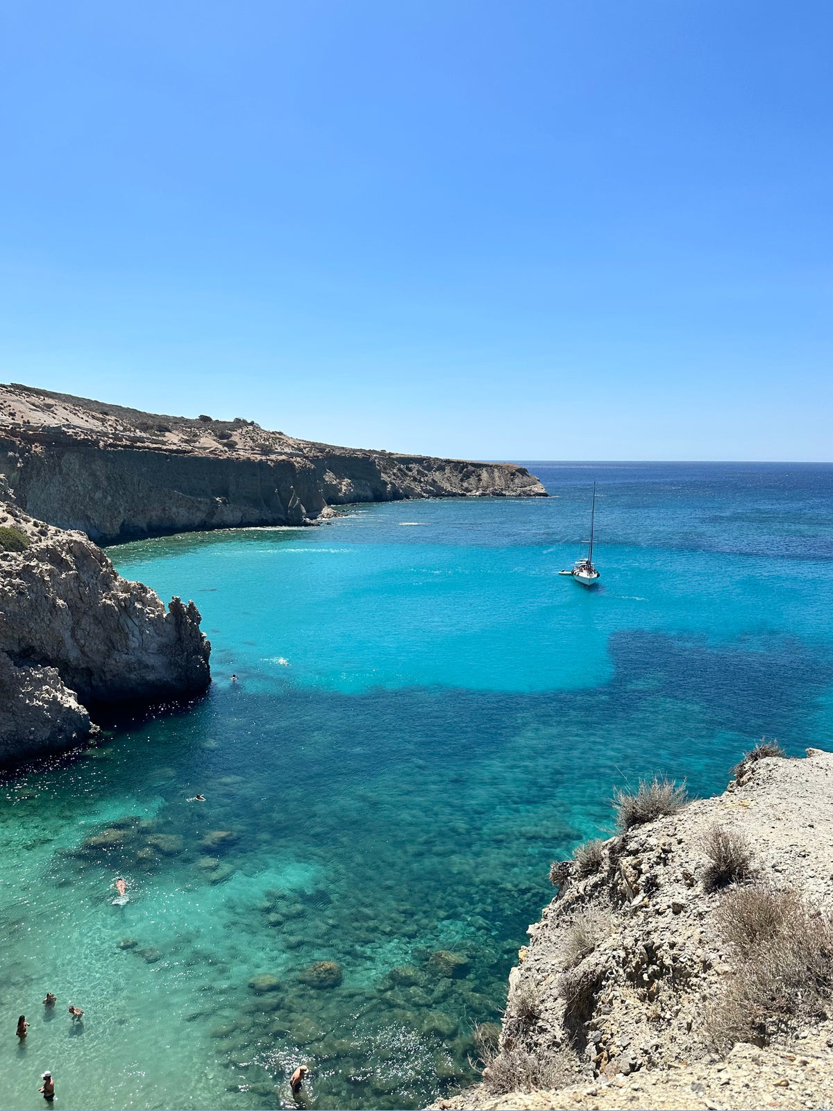 Sailing boat in a clear blue sea with rocky cliffs and people swimming.