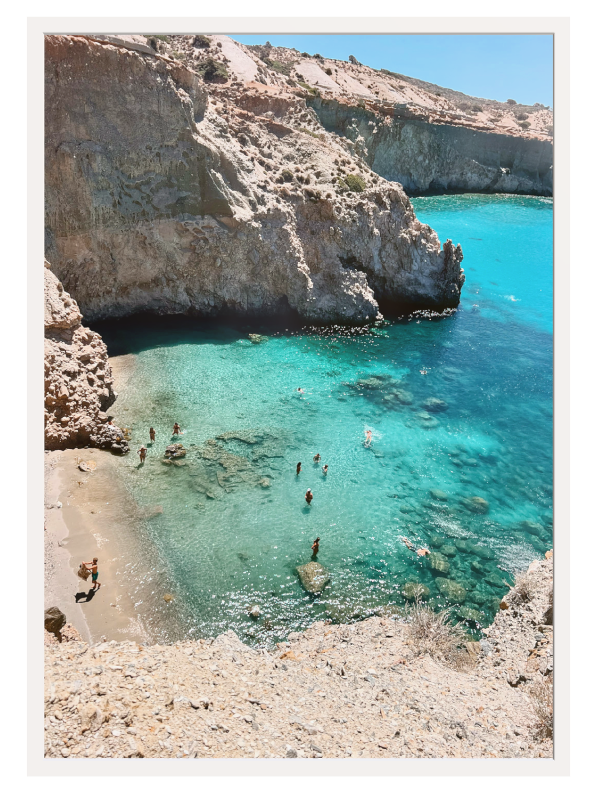 Cove with clear blue water and rocky cliffs, people swimming and sunbathing.
