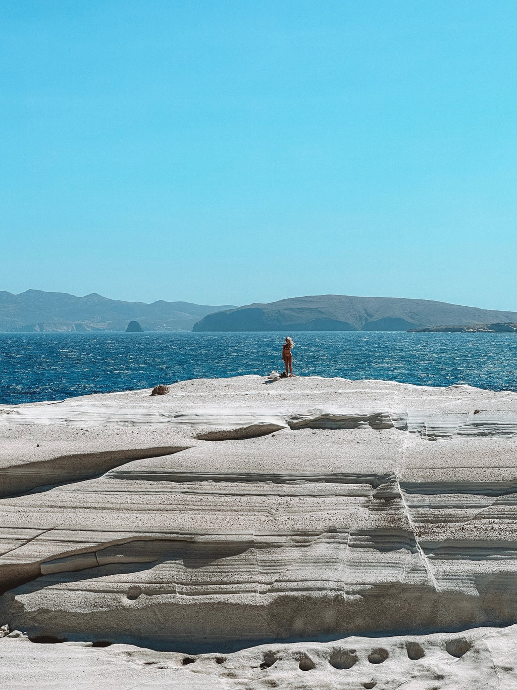 Person standing on a rocky outcrop overlooking the ocean with mountains in the background.