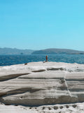 Person standing on a rocky outcrop overlooking the ocean with mountains in the background.