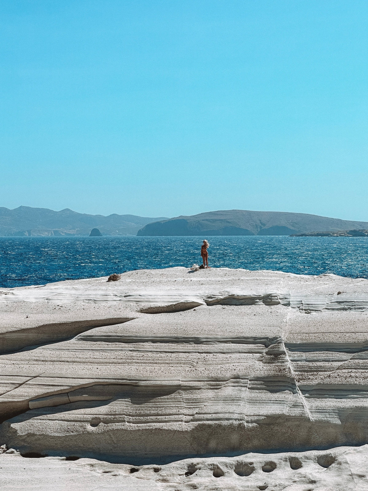 Person standing on a rocky outcrop overlooking the ocean with mountains in the background.