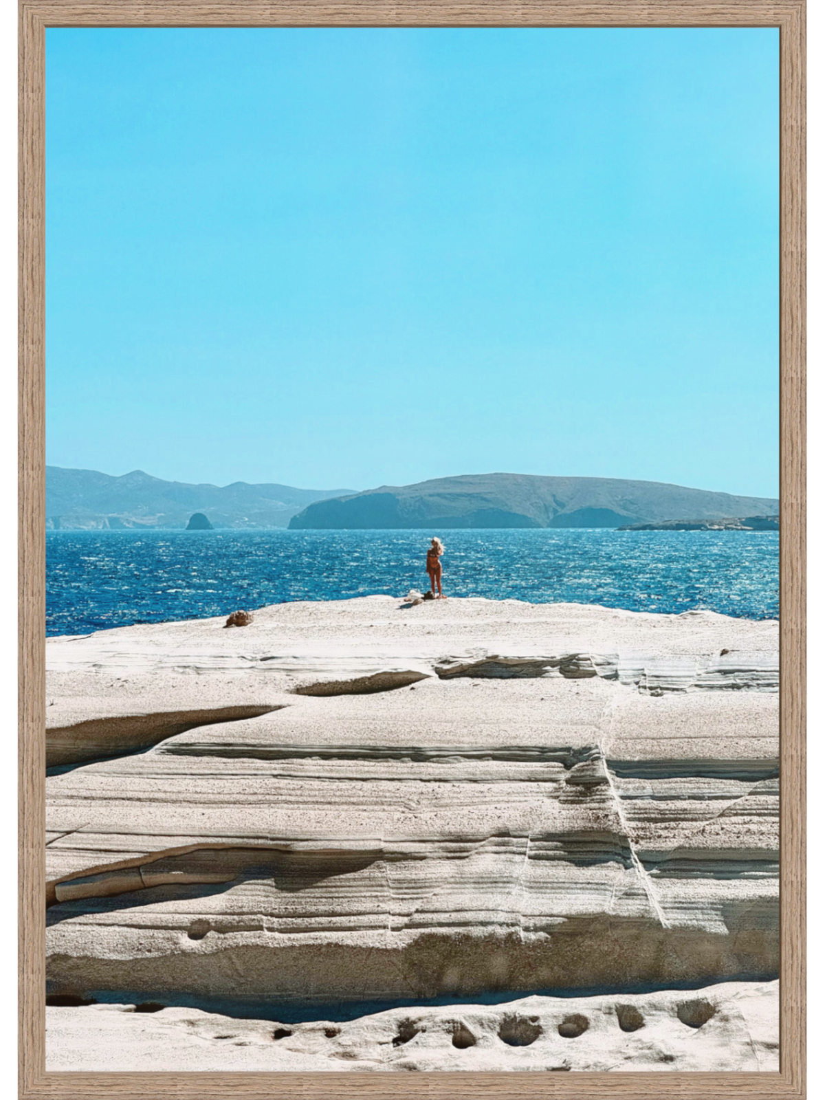 Framed photograph of a person on a rocky outcrop overlooking the ocean with mountains in the background.
