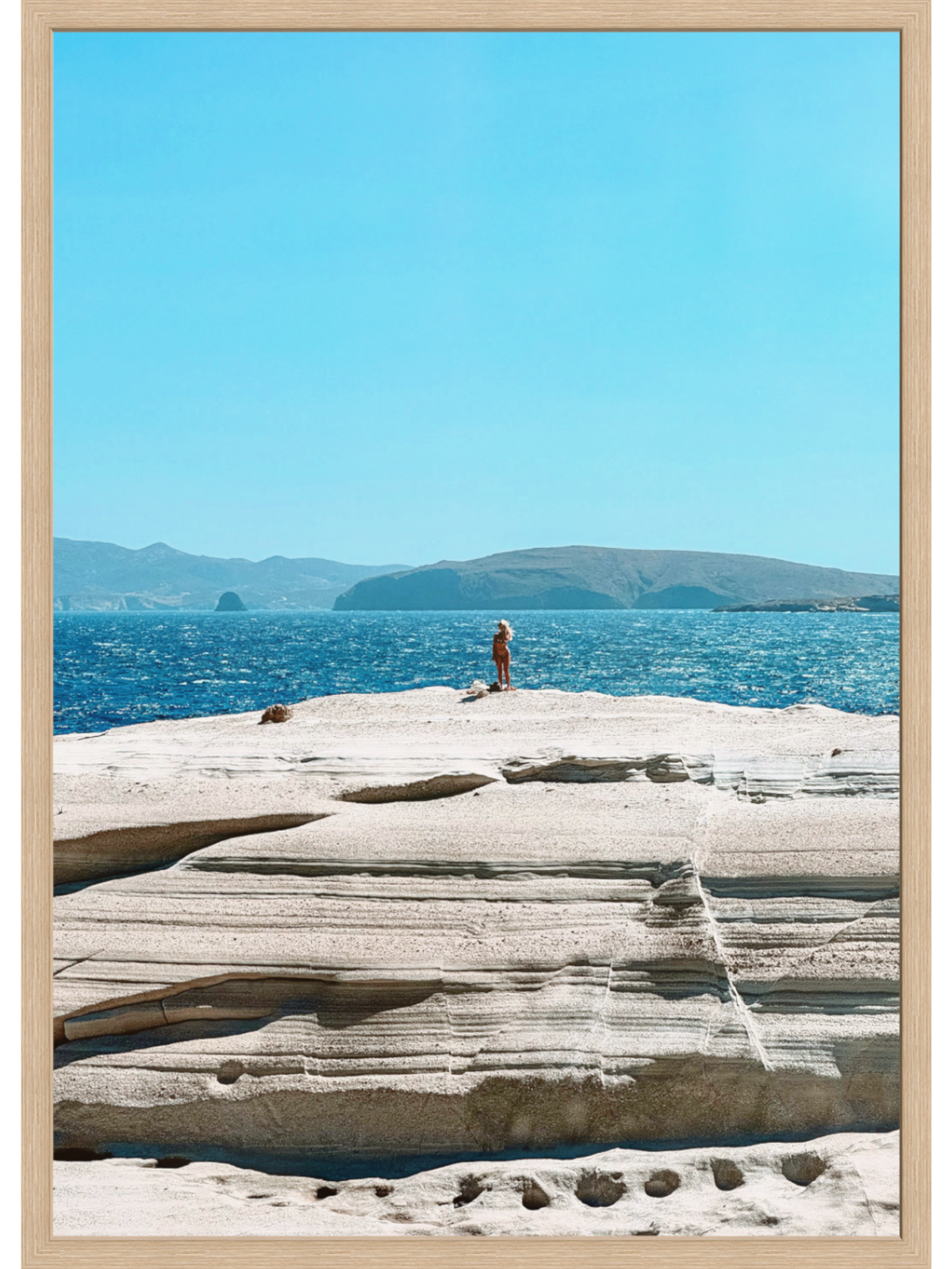 Person sitting on a rocky outcrop by the ocean with a clear blue sky.