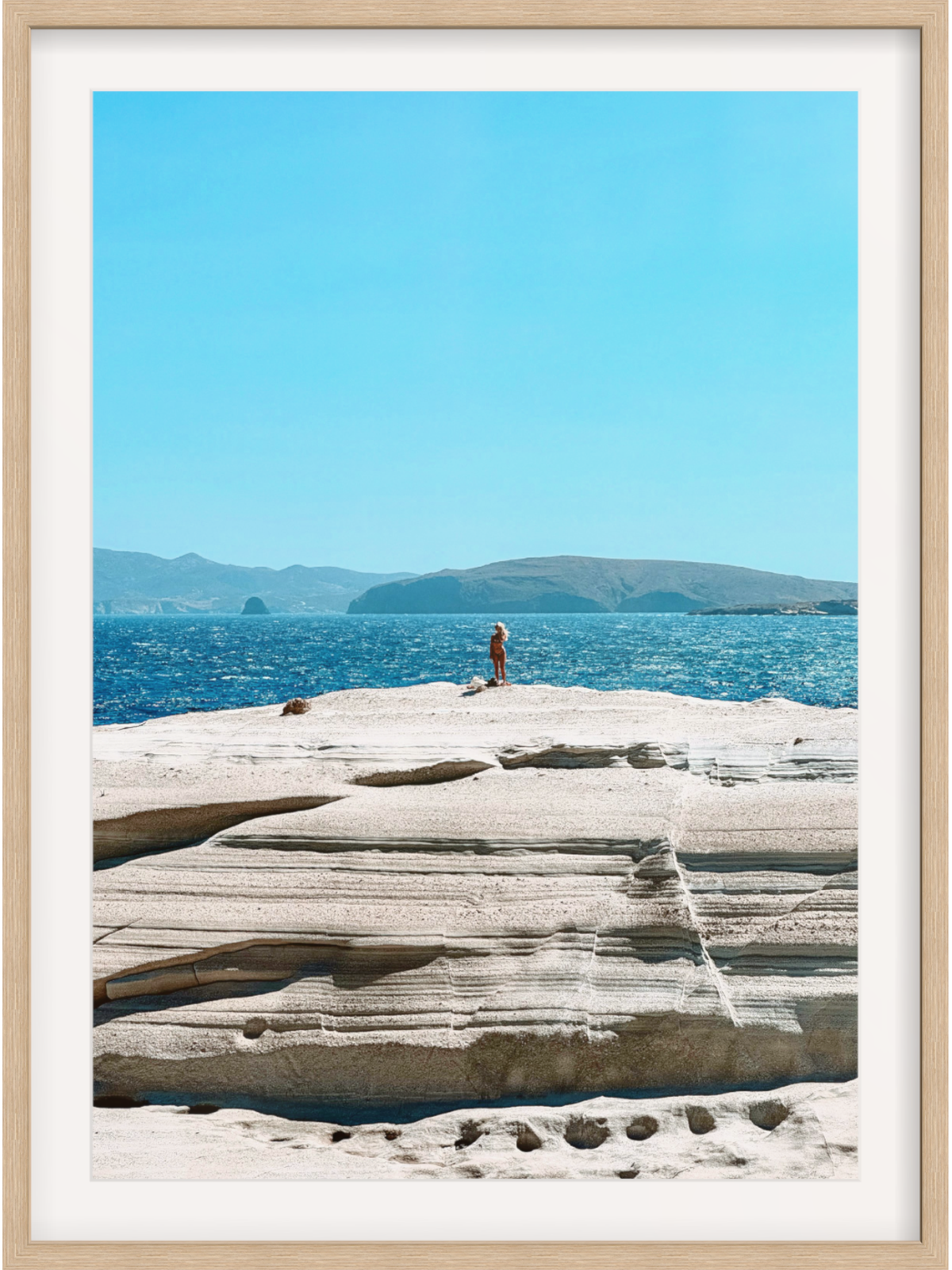 Person standing on a rocky outcrop with blue water and sky in the background, framed in a light wood border.