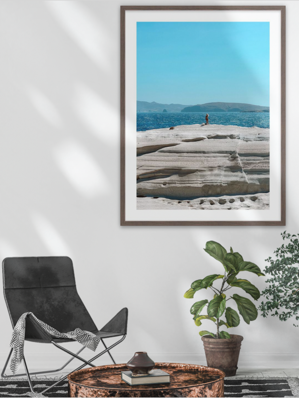 Framed artwork of a coastal scene with a person on rocks, displayed in a room with a black chair and plants.
