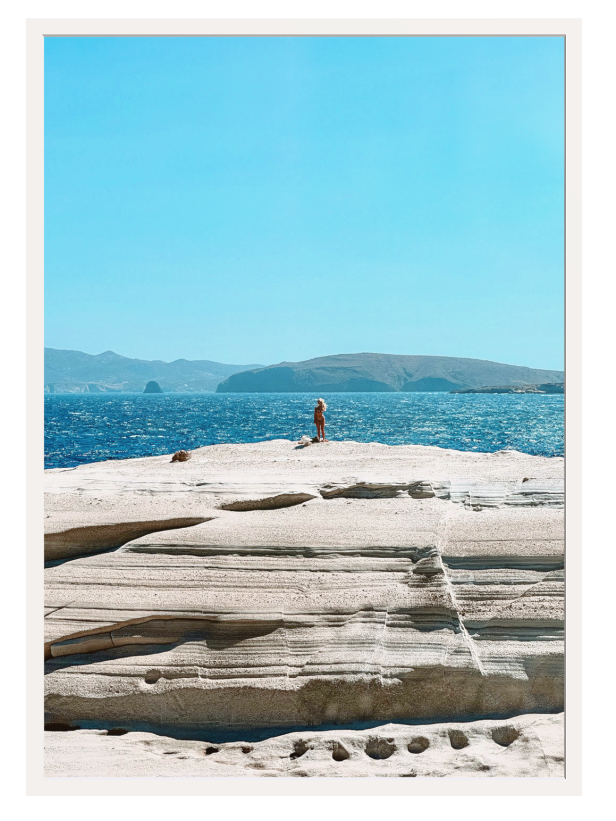 Person standing on a rocky outcrop with blue water and distant mountains.